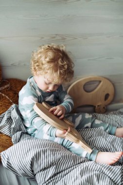 küçük kıvırcık bebeğim yatağa oturmuş ve ahşap bir oyuncakla oynamak mavi pijama