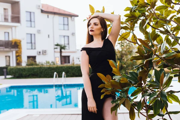 Attraente ragazza sexy in un abito nero in posa nel suo giardino accanto a una piscina blu — Foto Stock