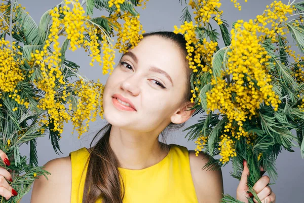 Hermosa joven en un vestido amarillo sostiene una mimosa amarilla fragante, sonríe y mira a la cámara — Foto de Stock