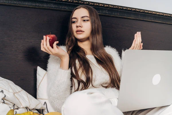 Linda chica de pelo largo sentada en la cama temprano en la mañana con su computadora portátil sosteniendo una manzana — Foto de Stock