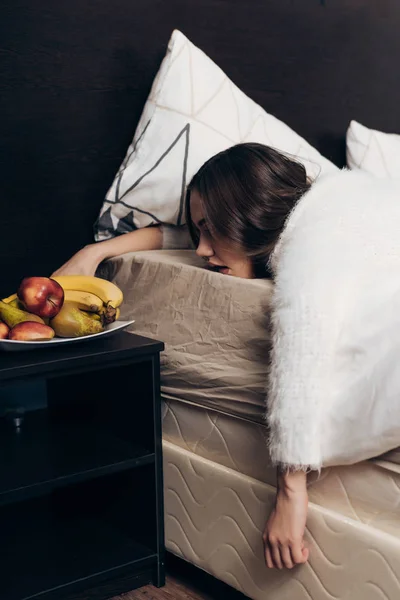 Jovem menina de pijama fica na cama de manhã cedo, um prato com frutas úteis para o café da manhã — Fotografia de Stock