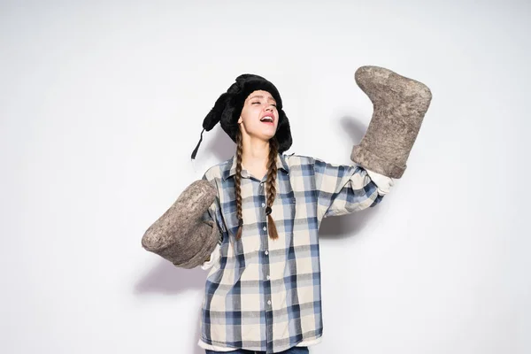 Divertida chica rusa joven con coletas, en un sombrero de piel caliente sostiene botas de fieltro gris y disfruta del invierno — Foto de Stock