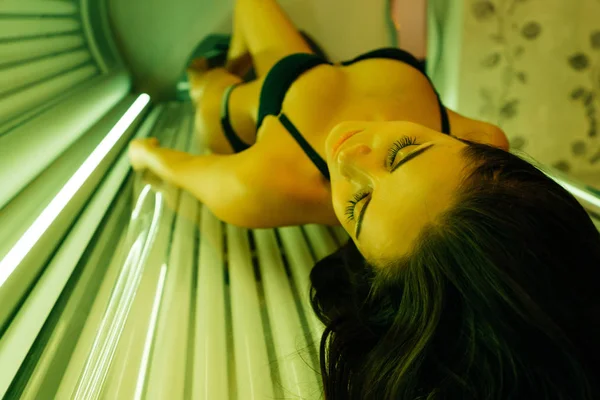 Una chica sexy delgada en un traje de baño negro se encuentra en una sala de sol y baños de sol bajo los rayos ultravioleta —  Fotos de Stock