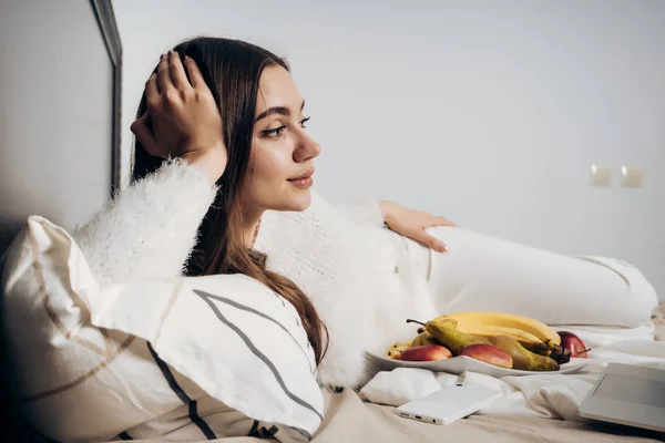 Linda chica de pelo largo en pijama blanco se encuentra temprano en la mañana en la cama, viendo una película y comiendo fruta — Foto de Stock