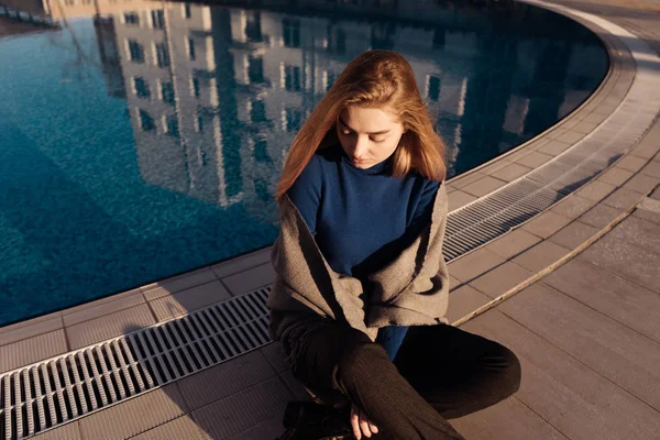 Krásná blondýnka ve stylový modrý svetr sedí u bazénu venku a odpočinek — Stock fotografie