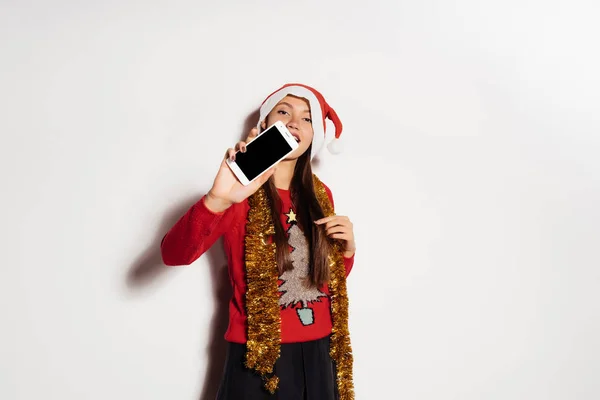Nettes junges Mädchen in rotem Neujahrspulli und Mütze wie der Weihnachtsmann feiert Neujahr und Weihnachten und hält ein Smartphone in der Hand — Stockfoto
