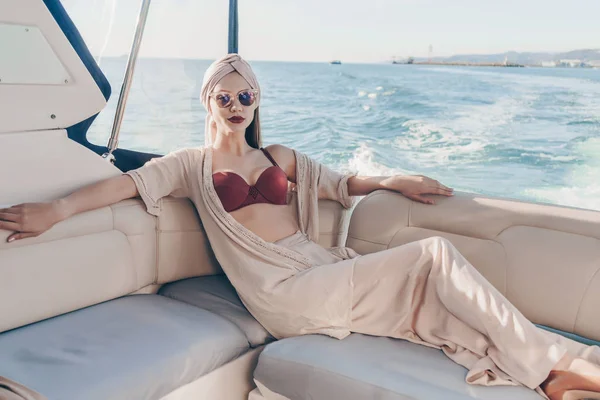 Una chica rica de lujo con un pañuelo en la cabeza en su elegante ropa de verano se relaja en su yate blanco, un viaje por el mar —  Fotos de Stock