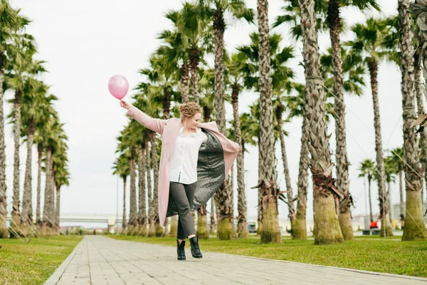 Een grappige stijlvolle vrouw met een lange roze vacht loopt het park naast hoog groen palmen, houdt een roze ballon — Stockfoto
