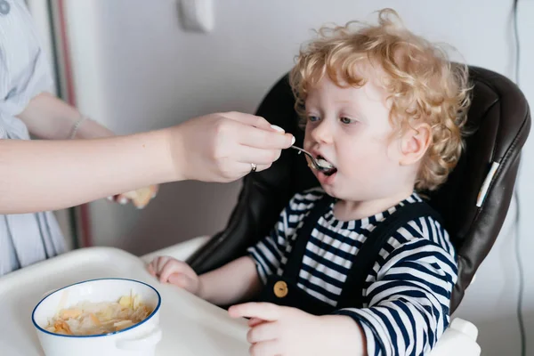 Заботливая молодая мать кормит своего маленького кудрявого сына здоровым супом, семейным завтраком на кухне — стоковое фото