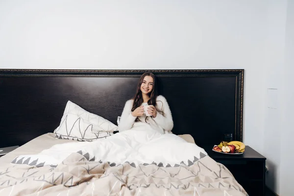 Linda chica joven en pijama sentado en la cama temprano en la mañana, buen humor, bebe café — Foto de Stock