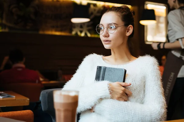 Милая юная студентка в белой куртке и очках, сидящая в кафе после школы, держа в руках блокнот и глядя в окно — стоковое фото