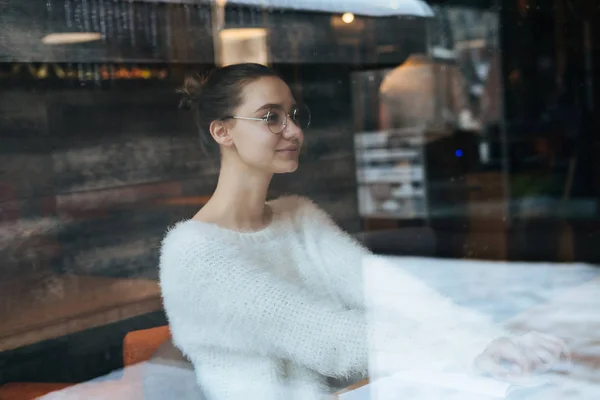 Красива молода дівчина-фрілансер у білому светрі та окулярах, сидить у кафе, думаючи про роботу, посміхаючись — стокове фото