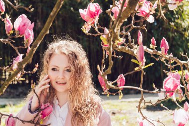 çekici kıvırcık genç kadın telefonda konuşurken Pembe Manolya yanında poz bahar park boyunca yürüyor