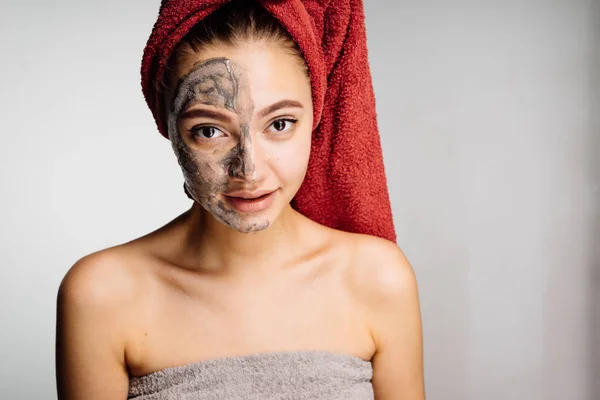 Una bella ragazza con un asciugamano rosso sulla testa ha applicato una maschera di argilla utile a metà del viso, una procedura spa — Foto Stock