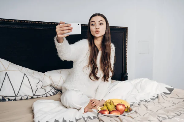 Una linda chica de pelo largo en pijama se sienta en la cama, se despierta temprano en la mañana, come fruta y hace selfie en su teléfono inteligente — Foto de Stock