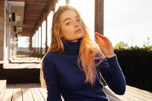 Bonito jovem loira menina em uma camisola azul na moda senta-se em um banco no parque ao sol da manhã, goza do calor e da primavera — Fotografia de Stock