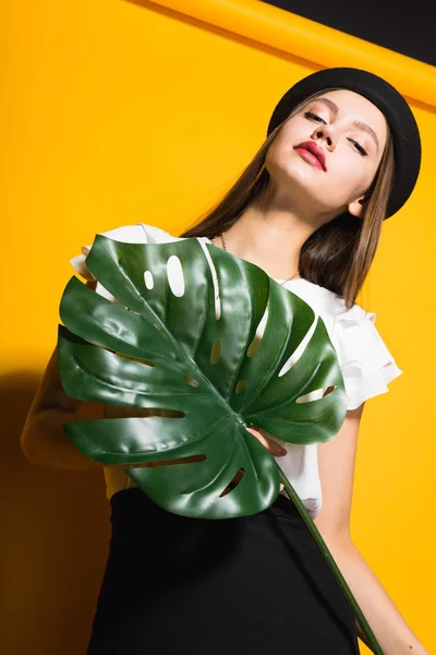 Jistý elegantní mladá dívka model v módním černém klobouku má zelený list a Pózování na žlutém podkladu — Stock fotografie
