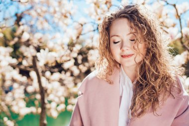 gülümseyerek, gözlerini kapsayan kokulu bir çiçek açan Manolya yanındaki bir parkta poz rahat kıvırcık genç kadın