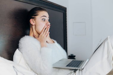 Pijama içinde genç sürpriz kız akşamları yatakta oturur ve ilginç film üstünde laptop görünüyor