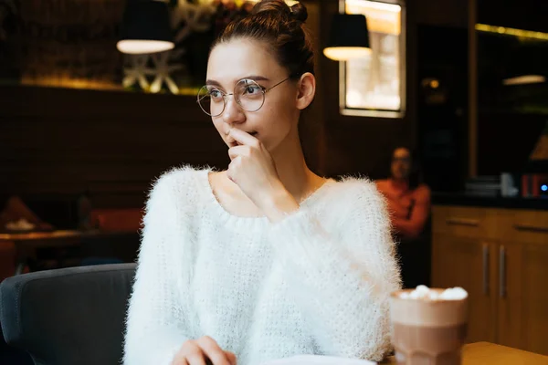 Красива молода дівчина студентка в білому светрі і окулярах сидить в затишному кафе після навчання в університеті і дивиться у вікно — стокове фото