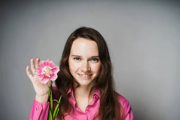 Bela jovem mulher em uma camisa rosa está segurando uma flor rosa perfumada, sorrindo e olhando para a câmera — Fotografia de Stock