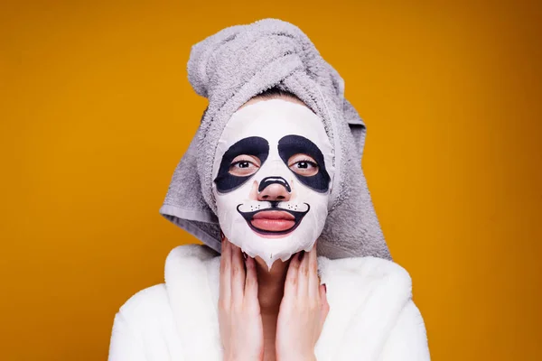 Een grappig meisje met een handdoek op haar hoofd en in een badjas behartigt de huid van het gezicht, op haar gezicht een gezichtsmasker met een gezicht van de panda — Stockfoto