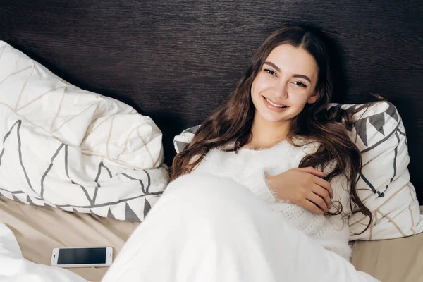 Schönes nettes Mädchen im weißen Pyjama liegt im Bett, ruht sich nach einem harten Arbeitstag aus und lächelt — Stockfoto