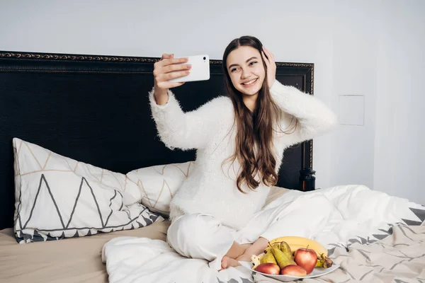 Mooi jong meisje in witte pyjama zit op een bed vroeg in de ochtend, maakt een dorp en glimlacht, fruit geserveerd — Stockfoto