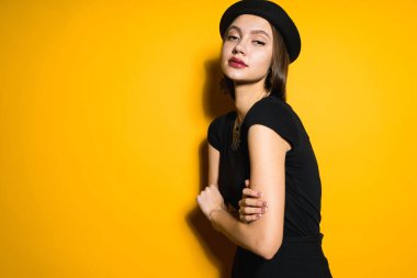 kendine güvenen lüks kız modeli siyah şık giysiler ve sarı zemin üzerine poz ve kameraya seyir şapka
