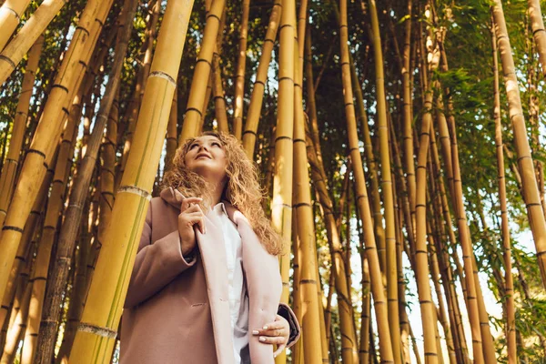 Stylish young woman in pink coat walking in park and posing next to tall bamboo trees — Stock Photo, Image