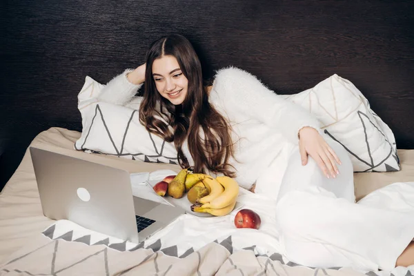 Gelukkig langhaar meisje in witte pyjama ligt in bed's avonds kijken naar een interessante film op haar laptop en het eten van fruit — Stockfoto