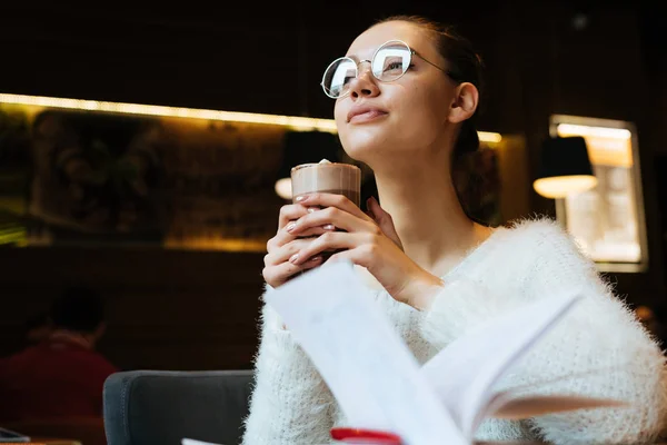 Красивая молодая девушка фрилансер в очках и белый свитер пьет вкусный латте в кафе и думает о работе — стоковое фото