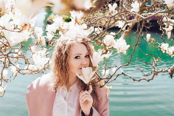 Curly elegant girl enjoys the scent of a blooming magnolia in a park next to a pond, poses — Stock Photo, Image