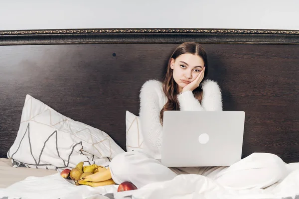 Tired young girl in pajamas sitting in bed and watching a boring movie on her laptop eating fruit — Stock Photo, Image