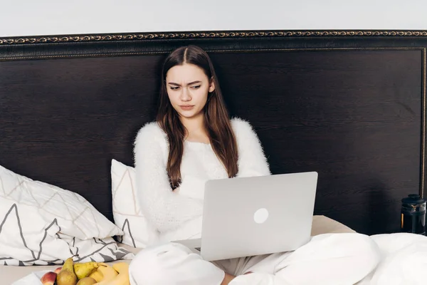 Joven insatisfecha en pijama blanco sentada en la cama por la noche y viendo una película de miedo en su computadora portátil — Foto de Stock