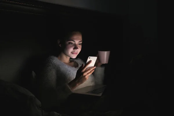 Una ragazza carina si siede su un letto al buio di notte, guardando un film su un computer portatile, bevendo tè e tenendo in mano uno smartphone — Foto Stock