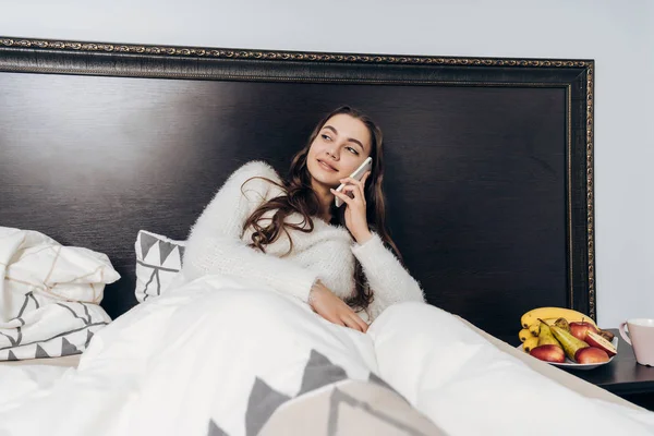 Bonito jovem menina em pijama branco encontra-se na cama no início da manhã, falando ao telefone e sorrindo — Fotografia de Stock