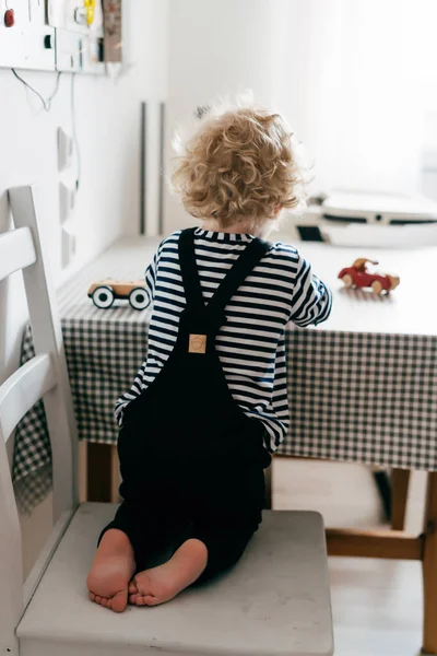 Kudrnatá malý chlapeček sedí na židli v kuchyni a hraje v hračkách — Stock fotografie