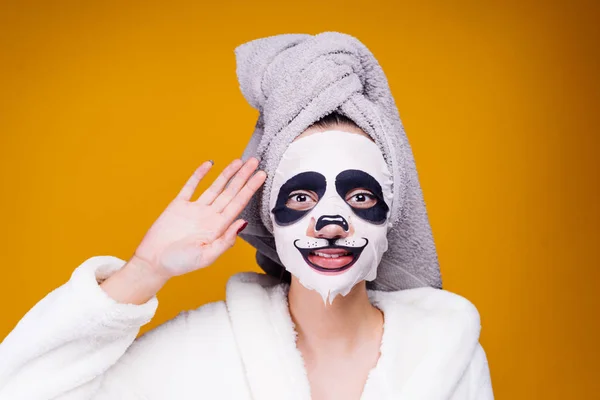 Een mooi jong meisje in een badjas en met een handdoek op haar hoofd zorg draagt voor zichzelf, op haar gezichtsmasker met een gezicht van de panda — Stockfoto