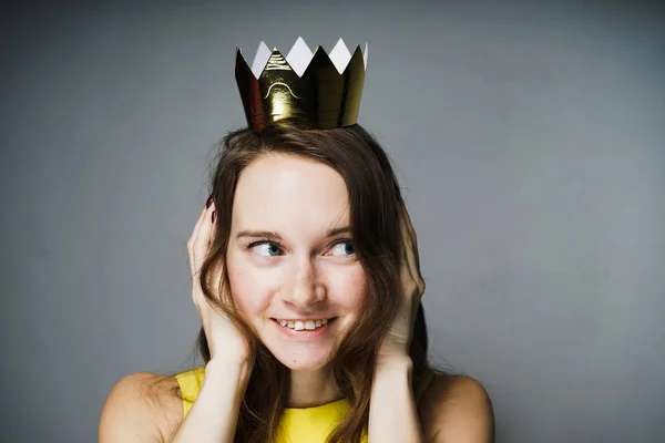 Divertida joven con un vestido amarillo sonríe, cubriéndose las orejas con las manos, sobre su cabeza una corona dorada — Foto de Stock