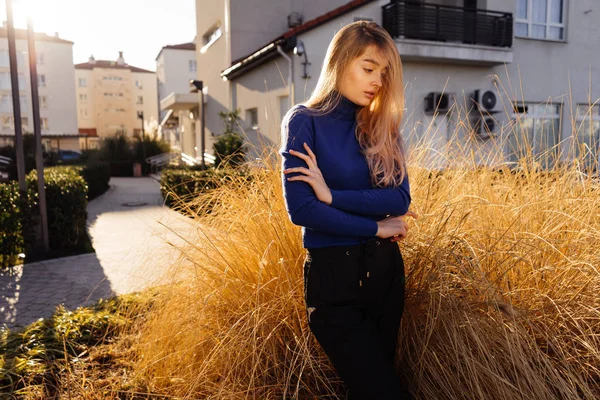 Menina loira elegante em suéter azul posando nas ruas da cidade ao sol — Fotografia de Stock