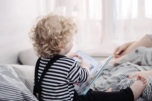 Lindo niño de pelo rizado en un traje de niños está sentado en la cama y leyendo un libro — Foto de Stock