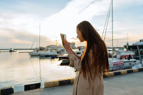 Stylowa dziewczyna długowłosy w beżowy płaszcz jest stojący w porcie morskim i wykonywanie zdjęć z seascape — Zdjęcie stockowe