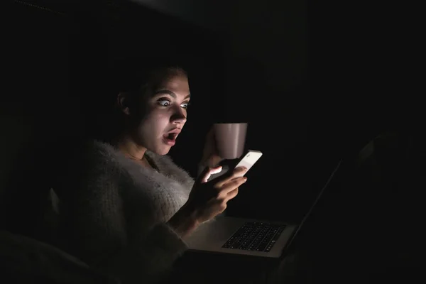 Giovane ragazza sorpresa seduta a letto la sera e guardando un film spaventoso sul suo computer portatile — Foto Stock