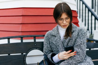 gri bir trendy ceket ve gözlük bir genç esmer şehir kız açık bir bankta oturur ve onu smartphone bir ileti yazar