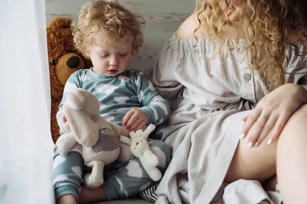 그의 엄마 옆에 앉아 그의 부드러운 장난감을가지고 노는 작은 곱슬 아기 — 스톡 사진