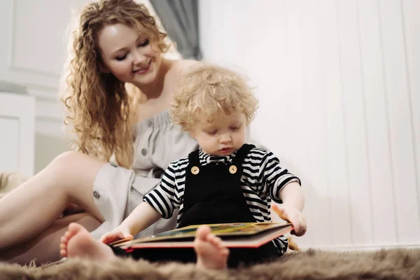 Feliz madre joven rizada sentada en el suelo con su hijo pequeño, leyendo un libro y divirtiéndose — Foto de Stock