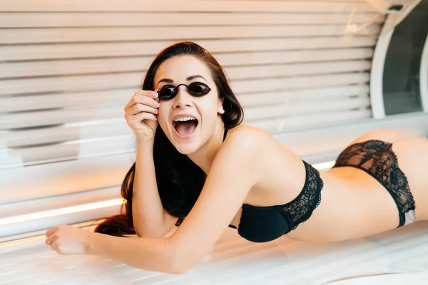 Menina morena esbelta feliz em maiô encontra-se na cama de sol horizontal e banhos de sol em óculos — Fotografia de Stock