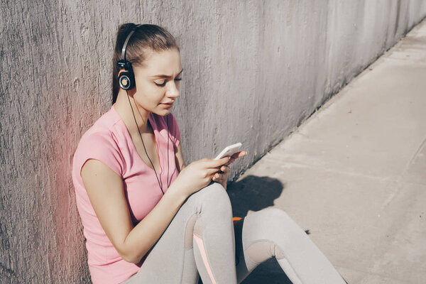 cute young girl in sportswear sitting on the ground outdoors, resting after a workout and listening to music on headphones