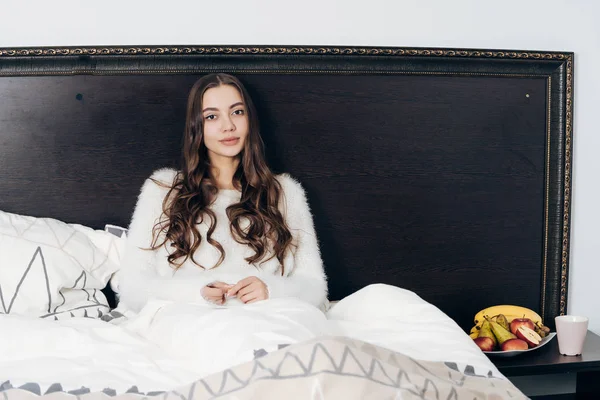 Schönes langhaariges Mädchen im weißen Pyjama sitzt im Bett, ruht sich nach einem harten Arbeitstag aus, will schlafen — Stockfoto